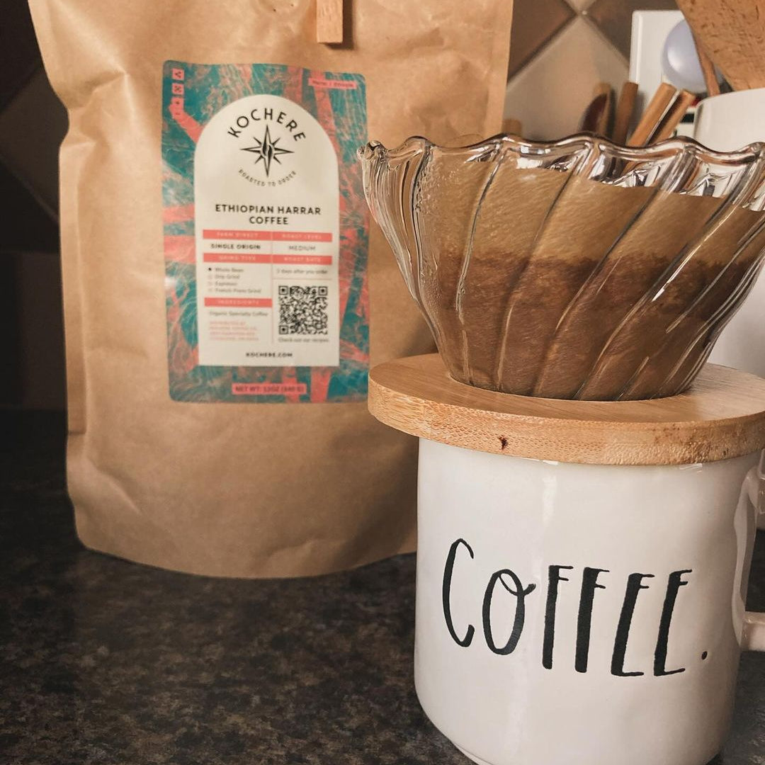 A pour-over coffee brewer sits on a white mug labeled “COFFEE” next to a bag of Kochere Ethiopian Harrar coffee on a kitchen counter.