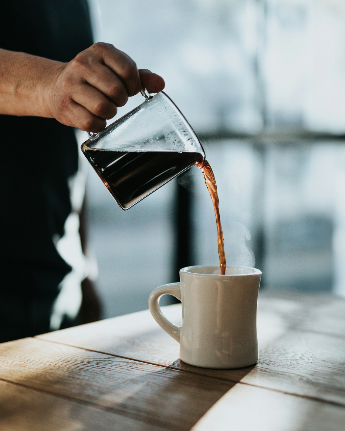 A person pours coffee from a glass carafe into a white mug on a wooden table, with sunlight coming through large windows in the background.