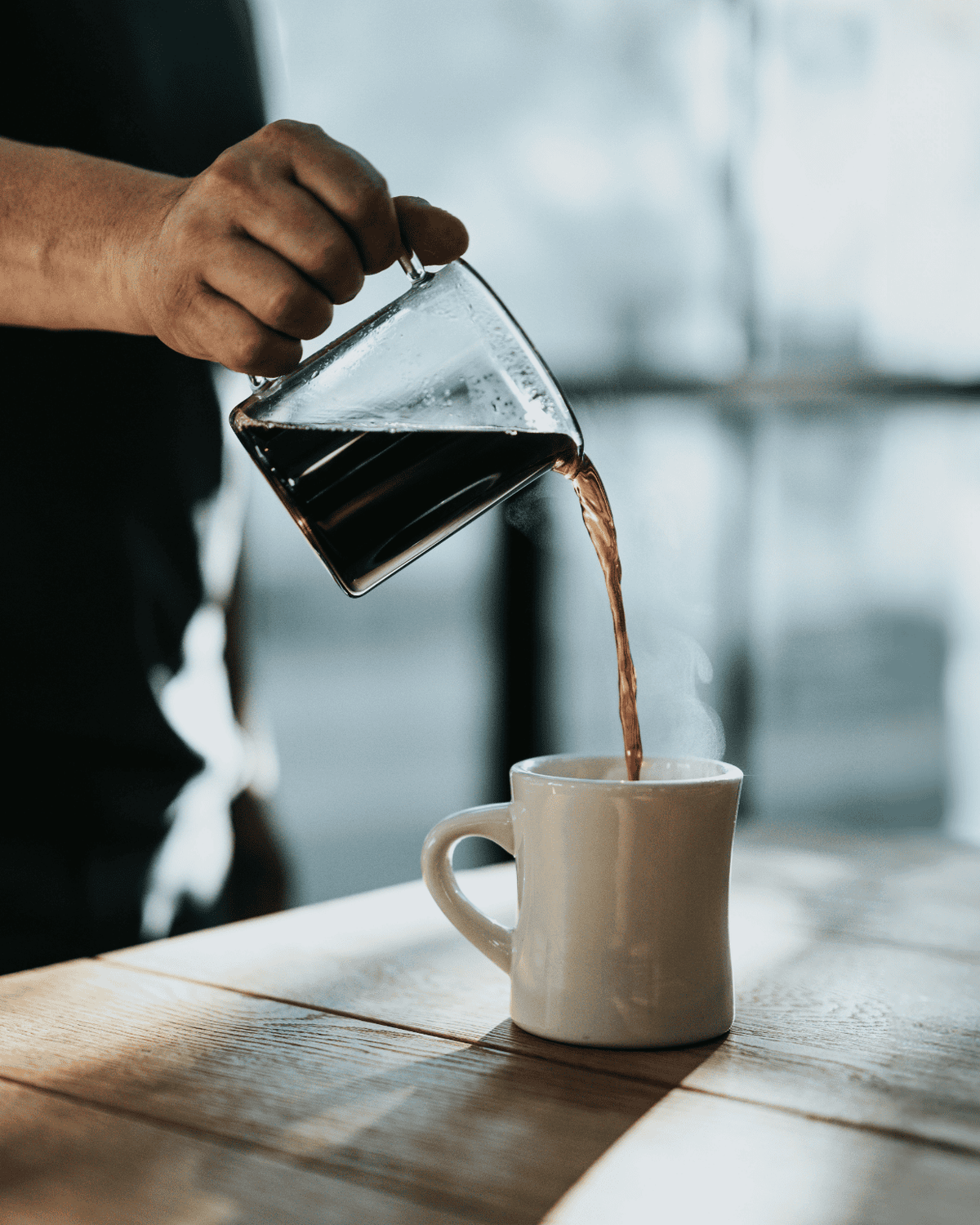 A person pours coffee from a glass carafe into a white mug on a wooden table, with sunlight coming through large windows in the background.