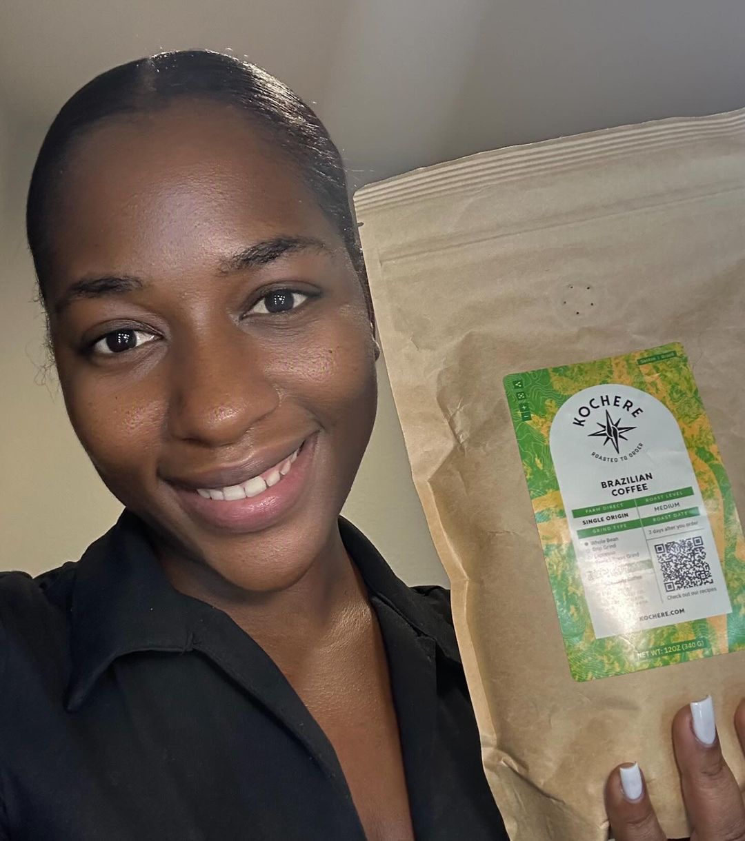 A person smiling and holding a brown bag of Nochere Brazilian Coffee in front of a neutral background.
