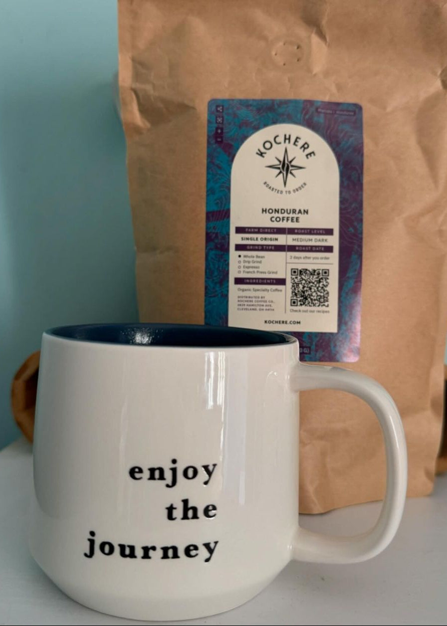 A white mug with "enjoy the journey" printed on it sits in front of a brown bag labeled "Kochere Honduran Coffee.