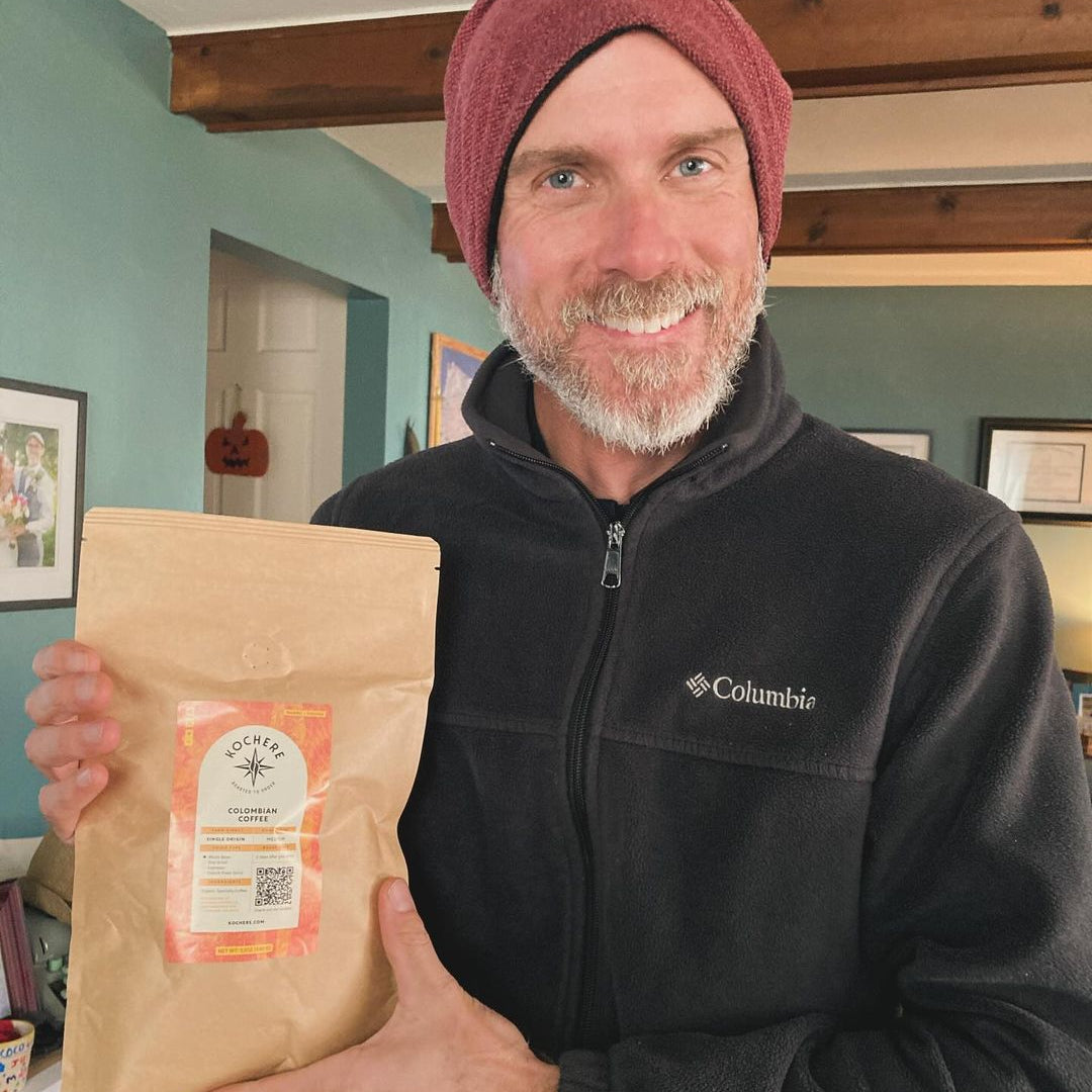 A man wearing a maroon beanie and black Columbia jacket smiles while holding a brown bag of coffee in a home setting.