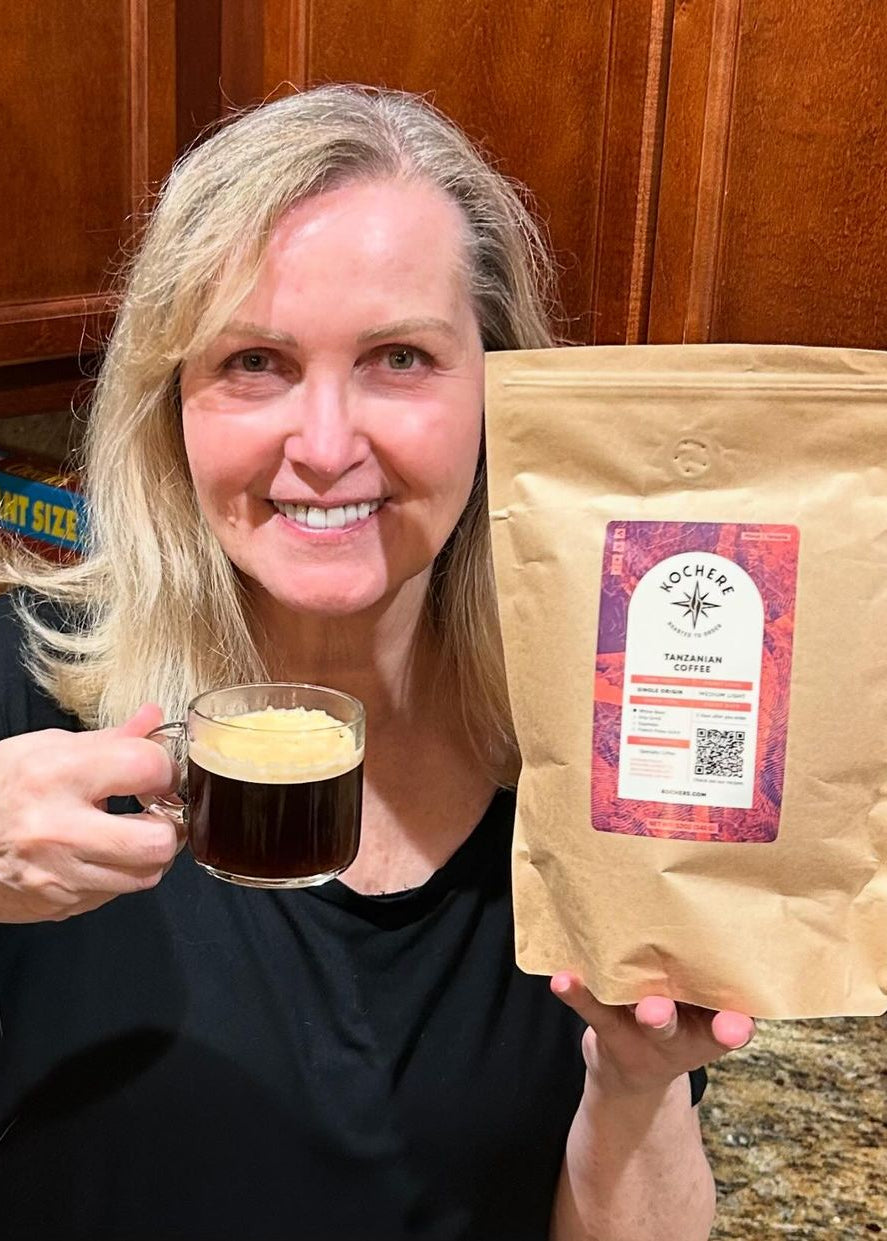 A woman smiles while holding a cup of coffee in one hand and a bag of Rocher Coffee Tanzanian Peaberry coffee beans in the other, in a kitchen setting.