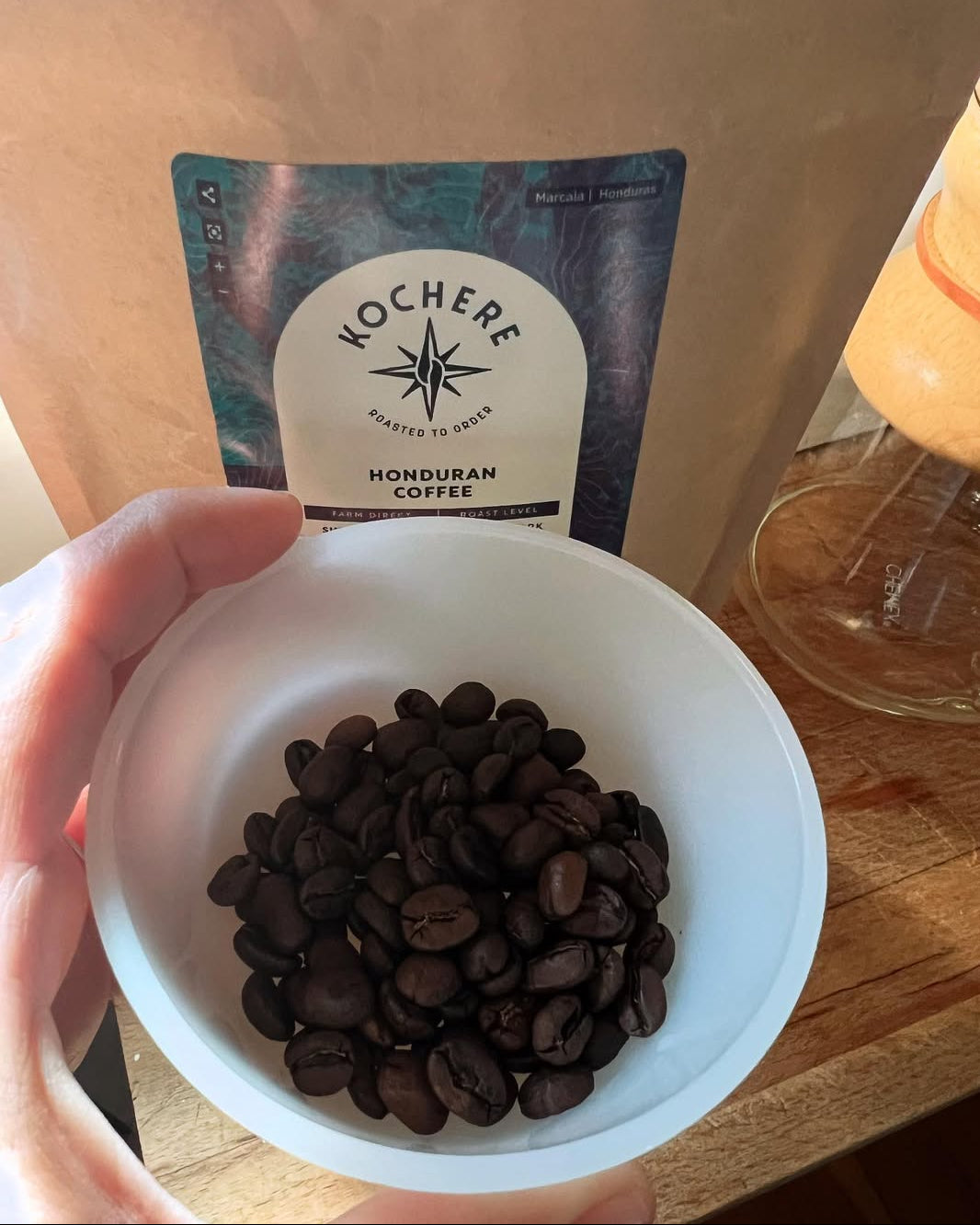 A hand holds a white bowl containing coffee beans in front of a bag labeled "Kochere Honduran Coffee" on a wooden surface.