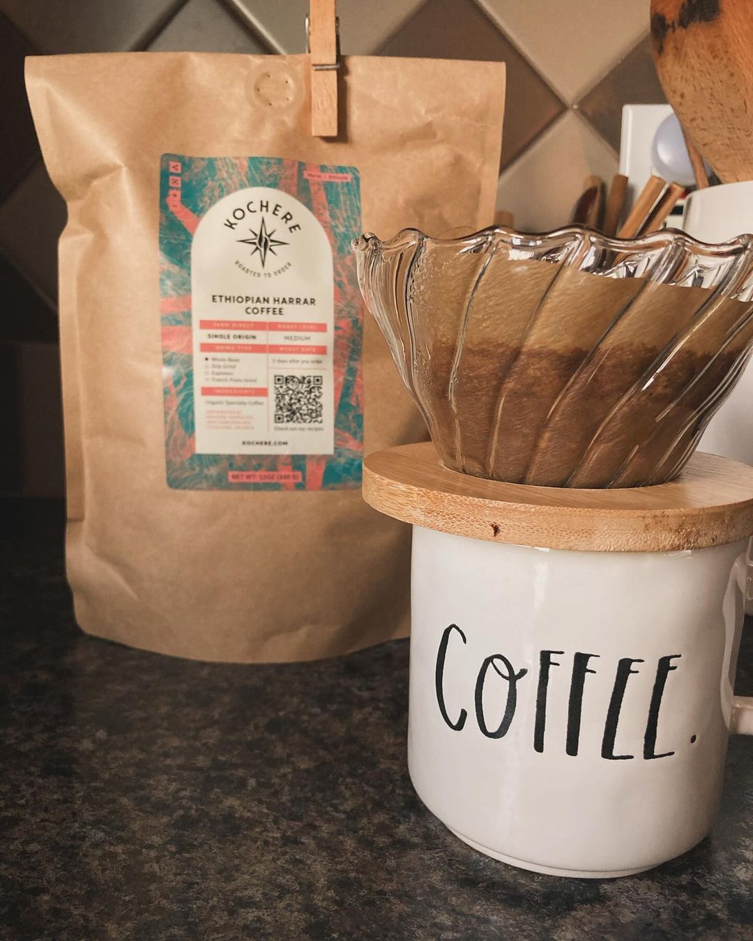 A bag of Ethiopian Harar coffee sits behind a glass pour-over on a white mug labeled "COFFEE" on a kitchen counter.