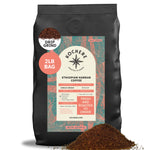 A 2lb bag of Kochere Coffee Ethiopian Harrar Natural Medium Roast by Kochere Coffee Company—single origin, organic, drip grind, fresh and roasted to order, with fruity blueberry and dark chocolate notes; beans and grounds spilled in front.