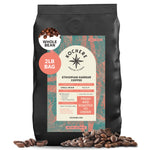 A 2lb bag of Kochere Coffee Ethiopian Harrar Natural Medium Roast by Kochere Coffee Company, featuring a colorful label, stands upright amid scattered coffee beans. Fruity blueberry and dark chocolate notes.