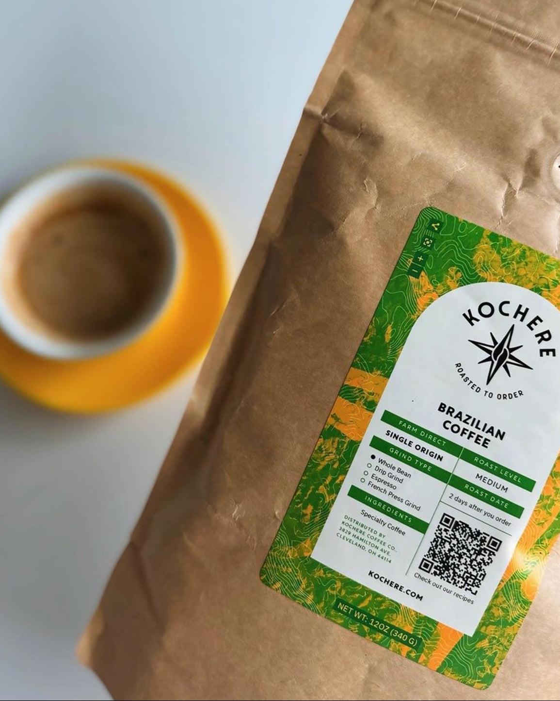 A bag of Kochere Brazilian coffee sits next to a cup of coffee in a yellow saucer. The coffee bag features a green label with product details and a QR code.
