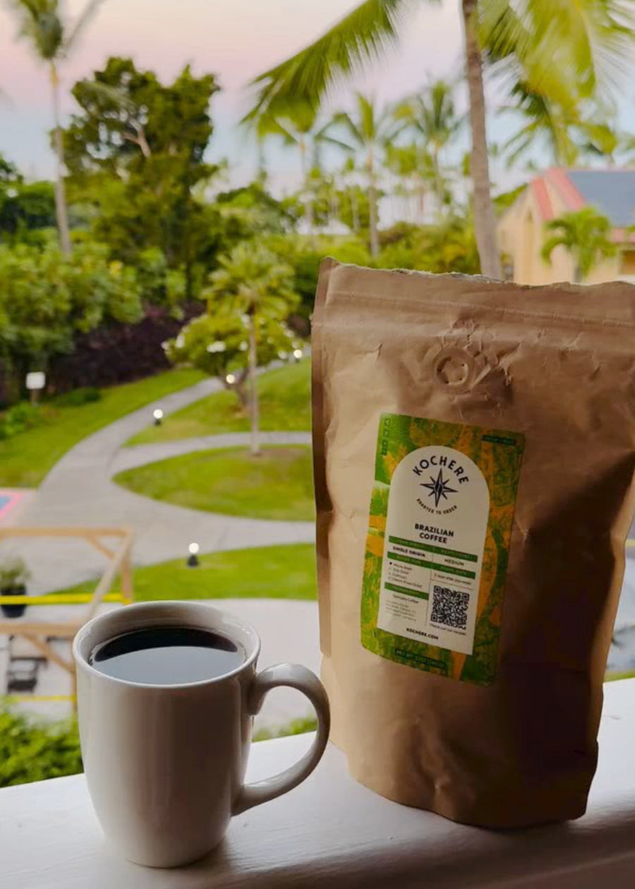 A white mug of black coffee and a brown bag of Kocher Brazilian coffee are placed on a balcony railing, with a tropical garden visible in the background.