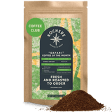 A brown bag labeled "Kochere Safari 'Coffee of the Month' Club" from Kochere Coffee Company sits with ground coffee in front, detailing roast level, single origin info, and club perks—ideal for your next fresh roasted coffee subscription.