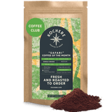 A brown bag labeled "Kochere Safari 'Coffee of the Month' Club - Fresh Roasted Single Origin Subscription" by Kochere Coffee Company sits beside ground coffee, featuring a green Coffee Club sticker—ideal for coffee subscription fans.