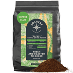 A black bag labeled "Kochere Safari 'Coffee of the Month' Club" by Kochere Coffee Company sits behind fresh ground coffee, highlighting single-origin, fresh-roasted details perfect for your next coffee subscription.[5LB][drip grind]