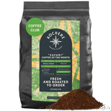 A black bag labeled "Kochere Safari 'Coffee of the Month' Club" by Kochere Coffee Company sits behind fresh ground coffee, highlighting single-origin, fresh-roasted details perfect for your next coffee subscription.