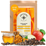 A 12 oz bag of Kochere Coffee Company’s Costa Rican Alajuela Medium Roast is displayed with coffee beans, a red apple, raisins, and caramel pieces, highlighting its sweet apple and raisin notes.[12 oz][Whole Bean]
