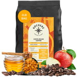 A 2lb bag of Kochere Coffee Company’s Costa Rican Alajuela Medium Roast—featuring sweet apple and raisin notes—is displayed with honey, raisins, coffee beans, and an apple artistically arranged in front of the package.[2LB][Whole Bean]