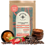 A 12 oz bag of Kochere Coffee Ethiopian Harrar Natural Medium Roast by Kochere Coffee Company stands upright, surrounded by coffee beans, strawberries, chocolate pieces, and a jar of chocolate spread.[12 oz][Whole Bean]