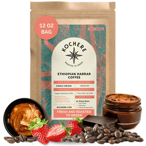 A 12 oz bag of Kochere Coffee Ethiopian Harrar Natural Medium Roast by Kochere Coffee Company stands upright, surrounded by coffee beans, strawberries, chocolate pieces, and a jar of chocolate spread.[12 oz][Whole Bean]
