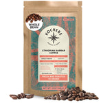 A 12oz bag of Kochere Coffee Ethiopian Harrar Natural Medium Roast by Kochere Coffee Company, with a colorful label, surrounded by scattered organic coffee beans. Features fruity blueberry and dark chocolate notes.