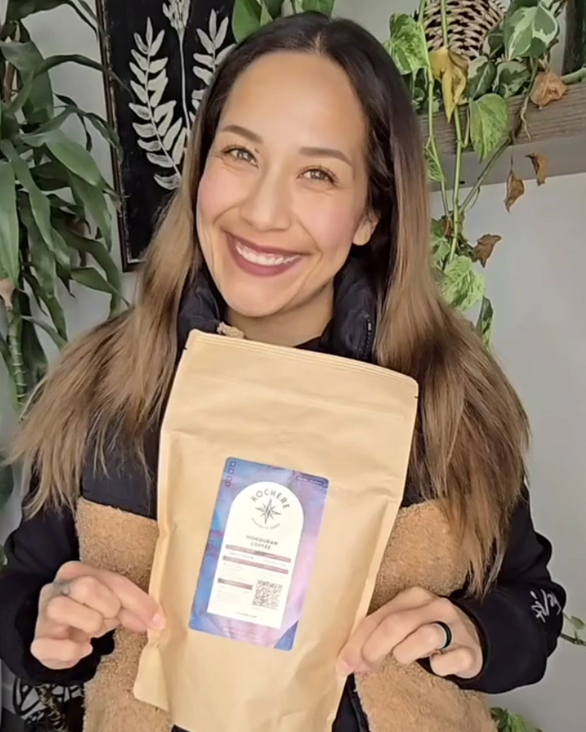 A woman standing indoors smiles and holds a brown bag of packaged product labeled "Rocky" with plants and decor in the background.