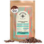 A 12oz bag of Kochere Coffee Ethiopian Sidamo Natural Medium-Light Roast by Kochere Coffee Company stands upright with scattered beans, featuring a green and orange design and noting its milk chocolate and fruity profile.