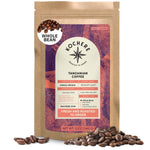 A 12 oz bag of Kochere Coffee Tanzanian Mbeya AB Medium-Light Roast, whole bean, with pear, floral, and jasmine notes stands upright amid scattered beans. The Kochere Coffee Company packaging shows brown, orange, and purple hues.