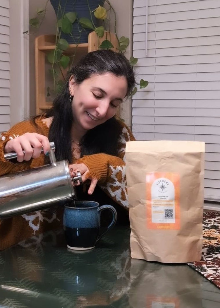 A woman pours coffee from a French press into a mug at a table. A bag of coffee beans is placed on the table next to her.