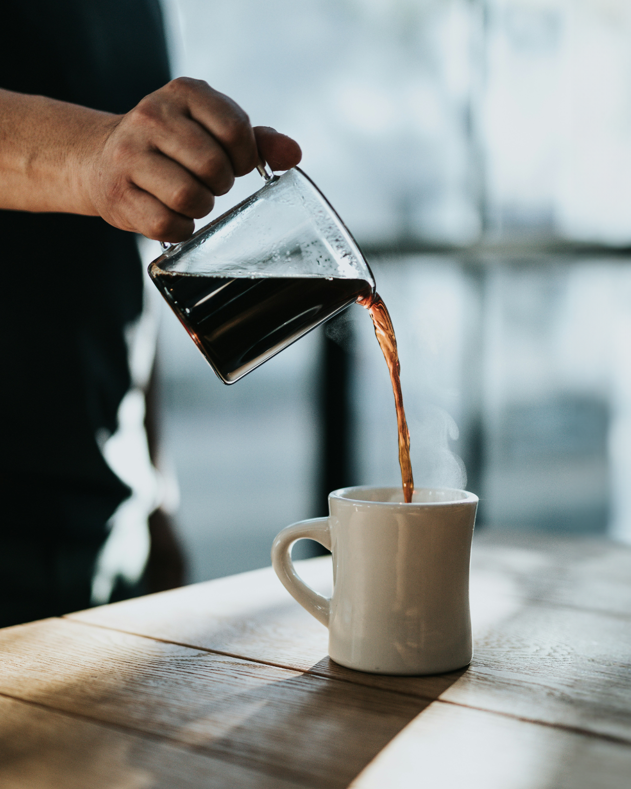 A person pours coffee from a glass carafe into a white mug on a wooden table, with sunlight coming through large windows in the background.