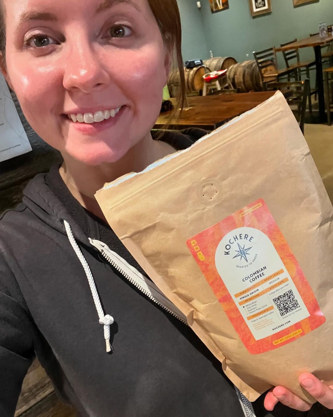 A woman in a hoodie holds a brown bag of coffee labeled "Kochere Colombian Coffee" in a cafe setting.