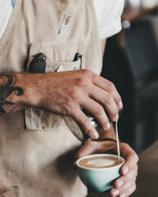 A person with a tattooed arm pours steamed milk into a cup of coffee, creating latte art, while wearing a beige apron.