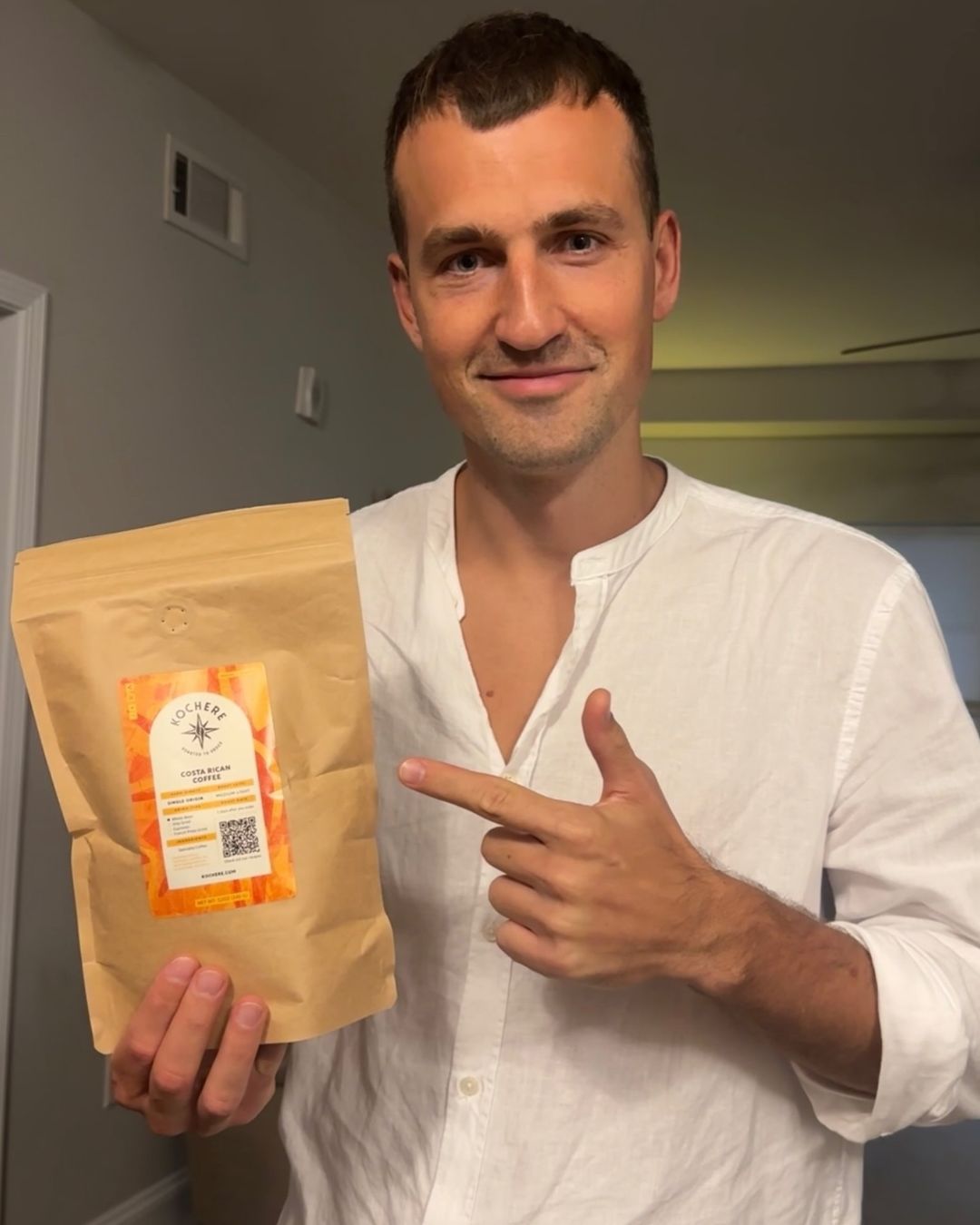 A man in a white shirt stands indoors, holding and pointing to a brown bag of packaged food or coffee with a visible label.