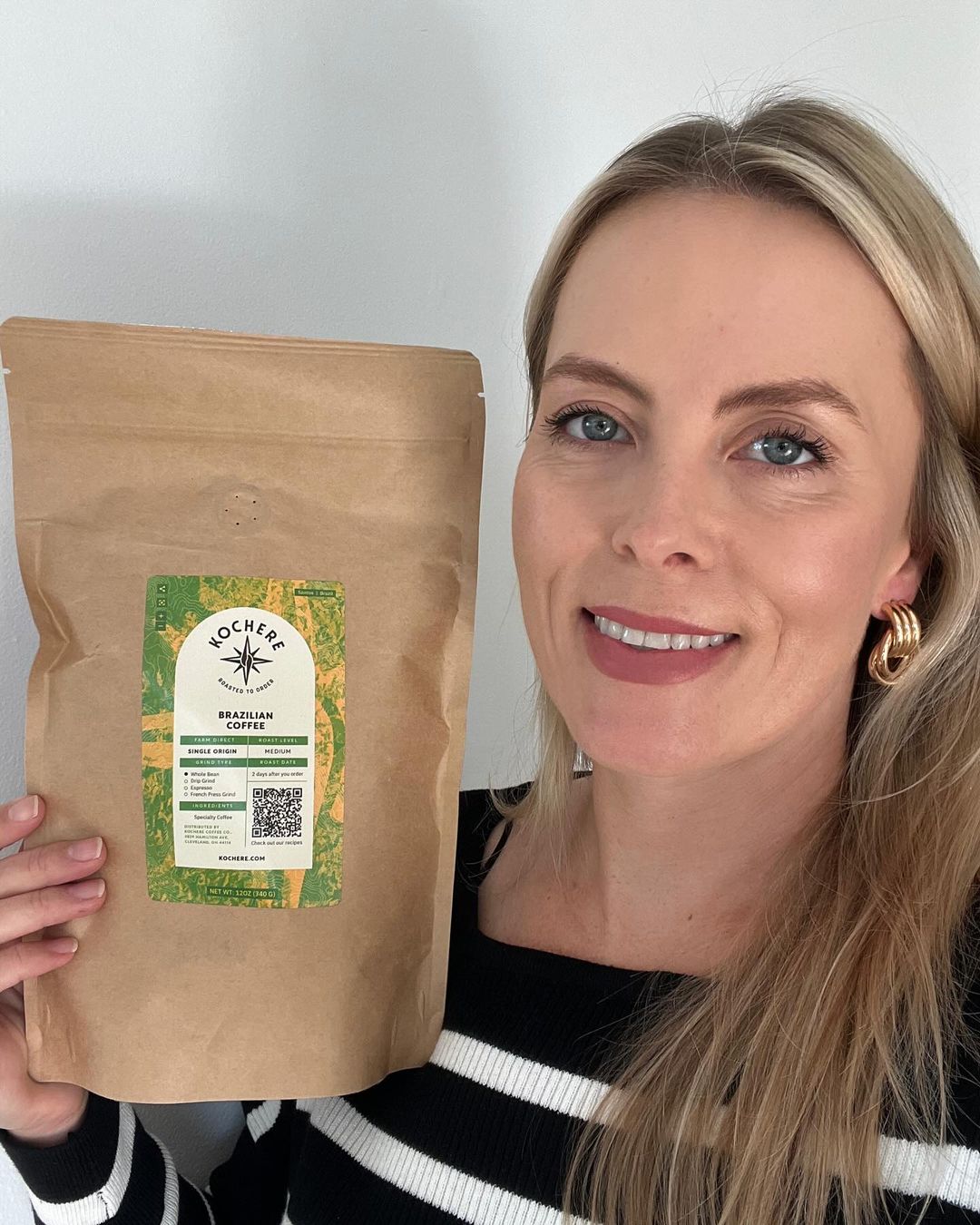 A woman holding a brown bag of Kocher Brazilian Coffee in front of a white wall.