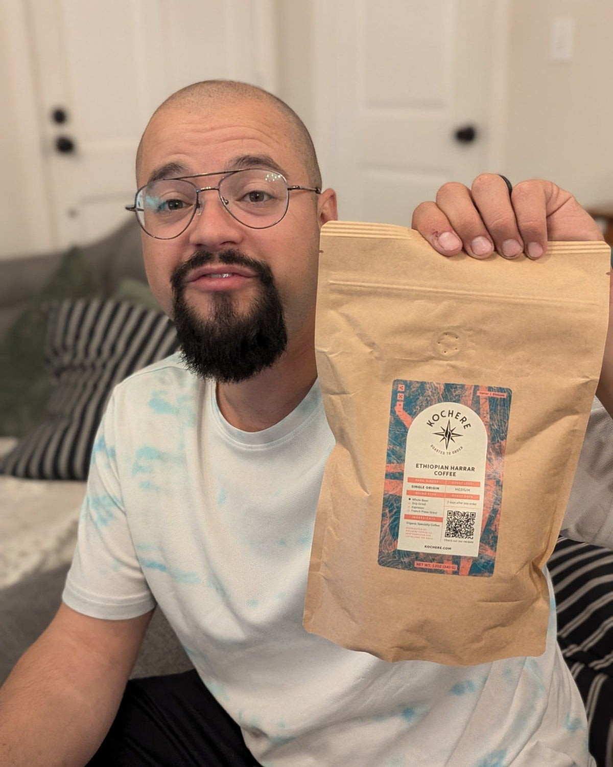 A man with glasses holds up a brown bag of Kocher Coffee labeled “Ethiopian Sidama.” He is seated indoors with a couch and pillows visible in the background.