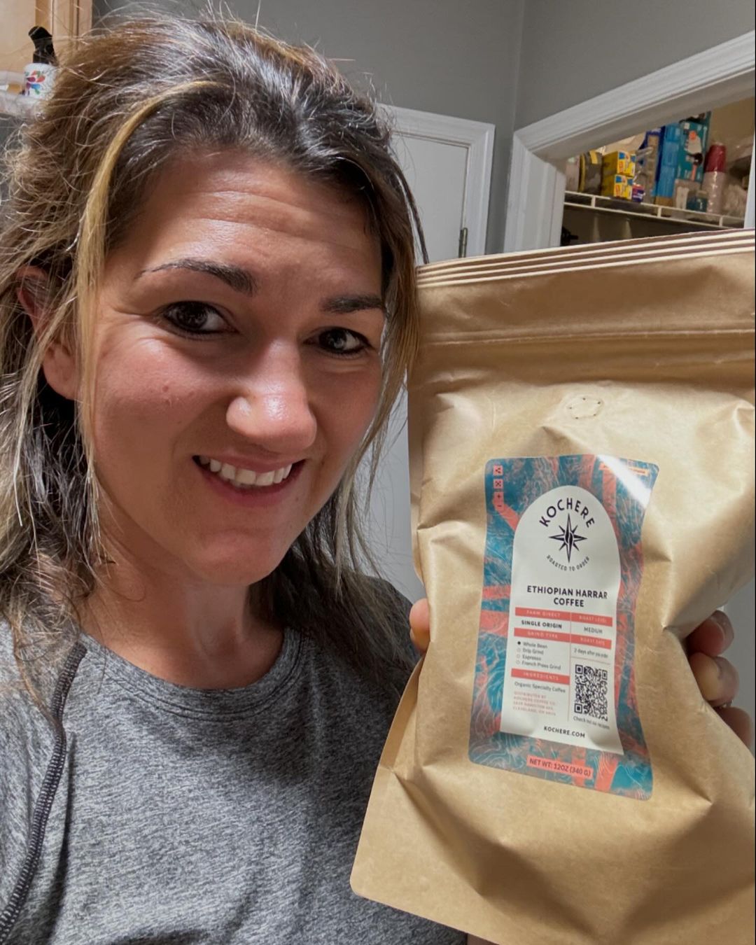 Person holding a large brown bag of Kocher Ethiopian Harar coffee in a kitchen, smiling at the camera.