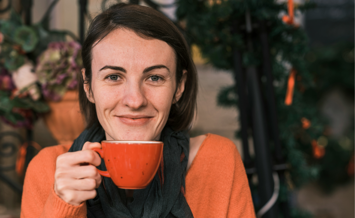 A person wearing an orange sweater and dark scarf smiles while holding a red mug, with blurred plants and decorations in the background.