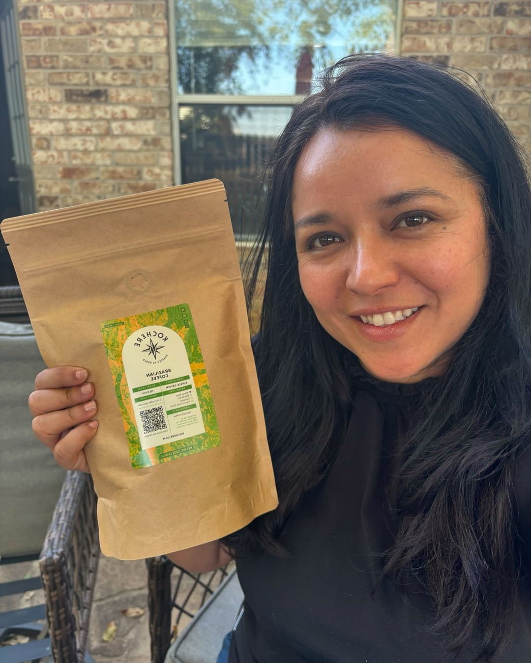 A woman holds up a brown bag of coffee with a green label while smiling at the camera outdoors.