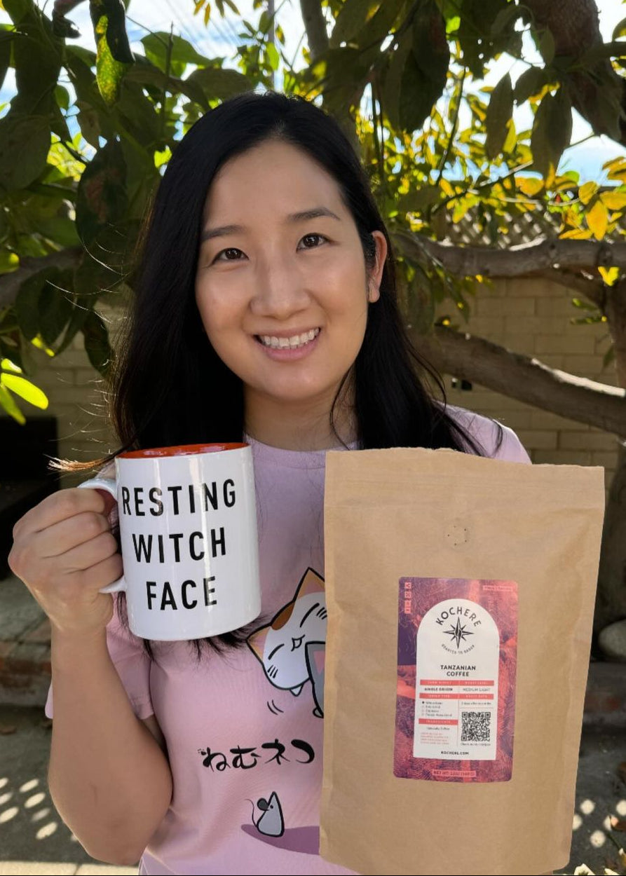 A woman smiles outdoors, holding a mug labeled "RESTING WITCH FACE" in one hand and a brown bag of coffee labeled "Kochere Ethiopia" in the other.