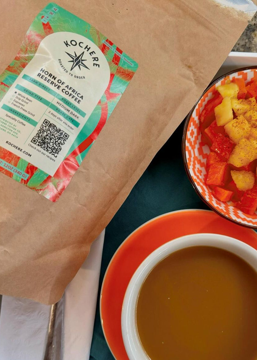 A bag of Kocherie Horn of Africa Reserve coffee sits beside a bowl of mixed fruit and a cup of coffee on an orange saucer.